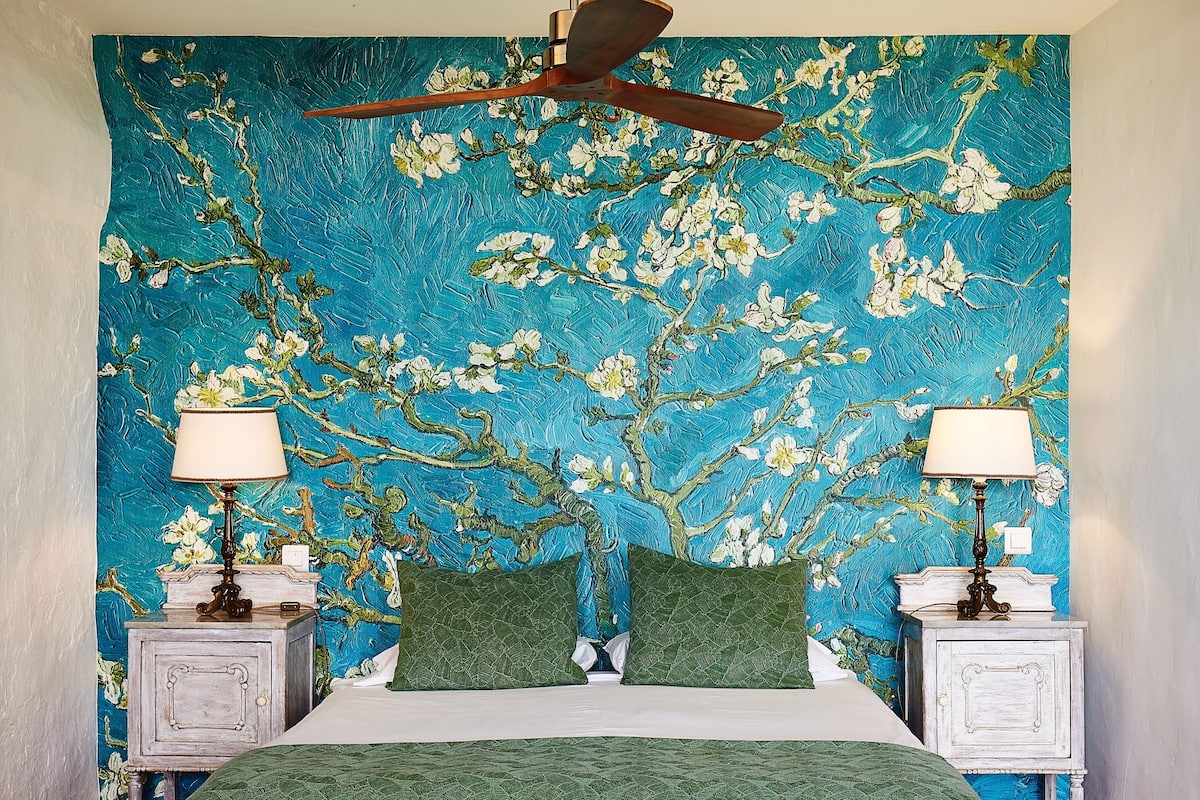 A bedroom with a large floral tapestry on the wall, a bed with green bedding, two bedside tables with lamps, and a ceiling fan.