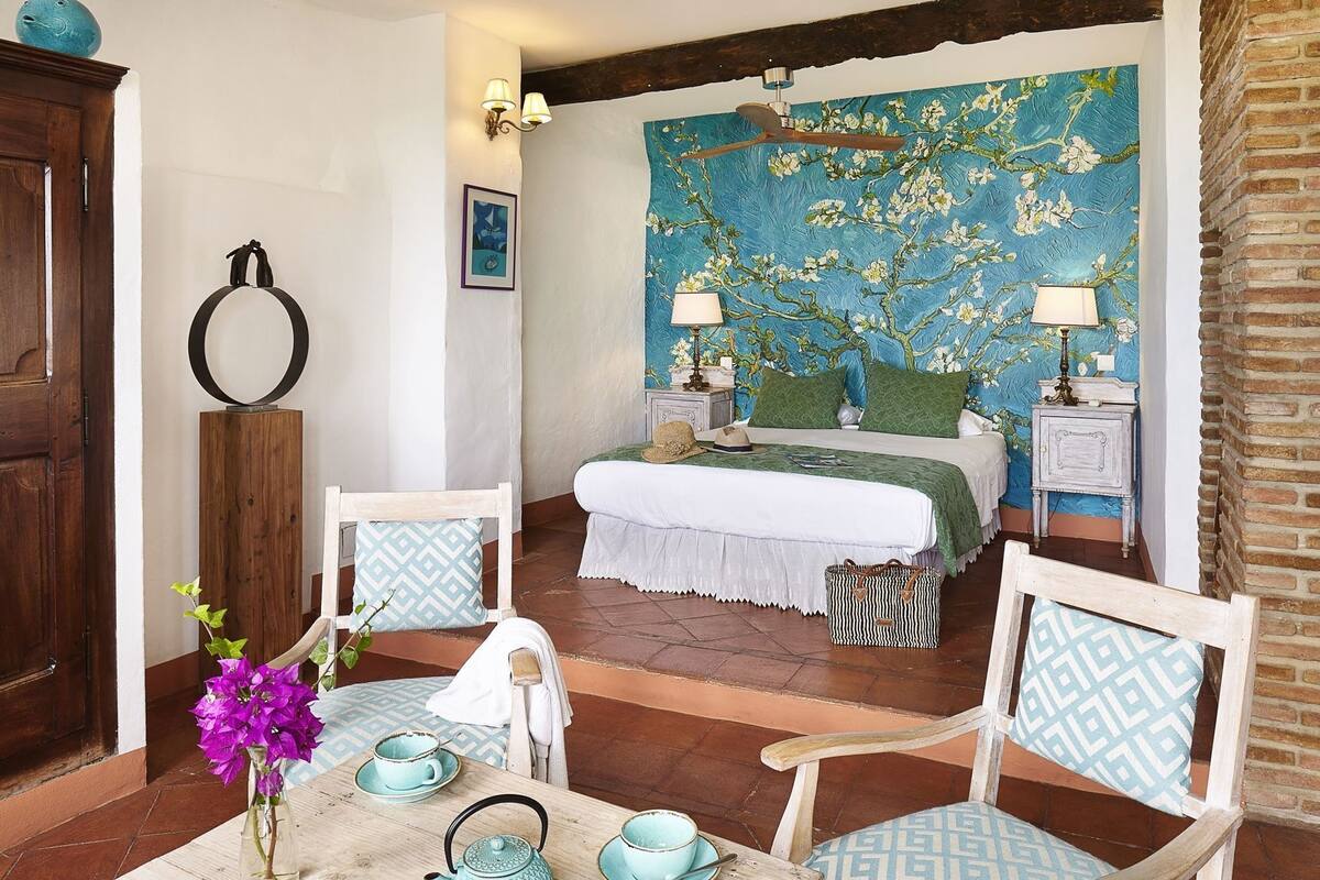 A bedroom with a bed, two bedside tables, a nightstand, a wooden chair, a table with a vase of flowers, and a wall with a floral mural.