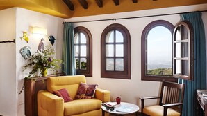 Un cuarto acogedor con un sofá amarillo, una silla de madera, una mesita y tres ventanas arqueadas que ofrecen vistas al paisaje.
