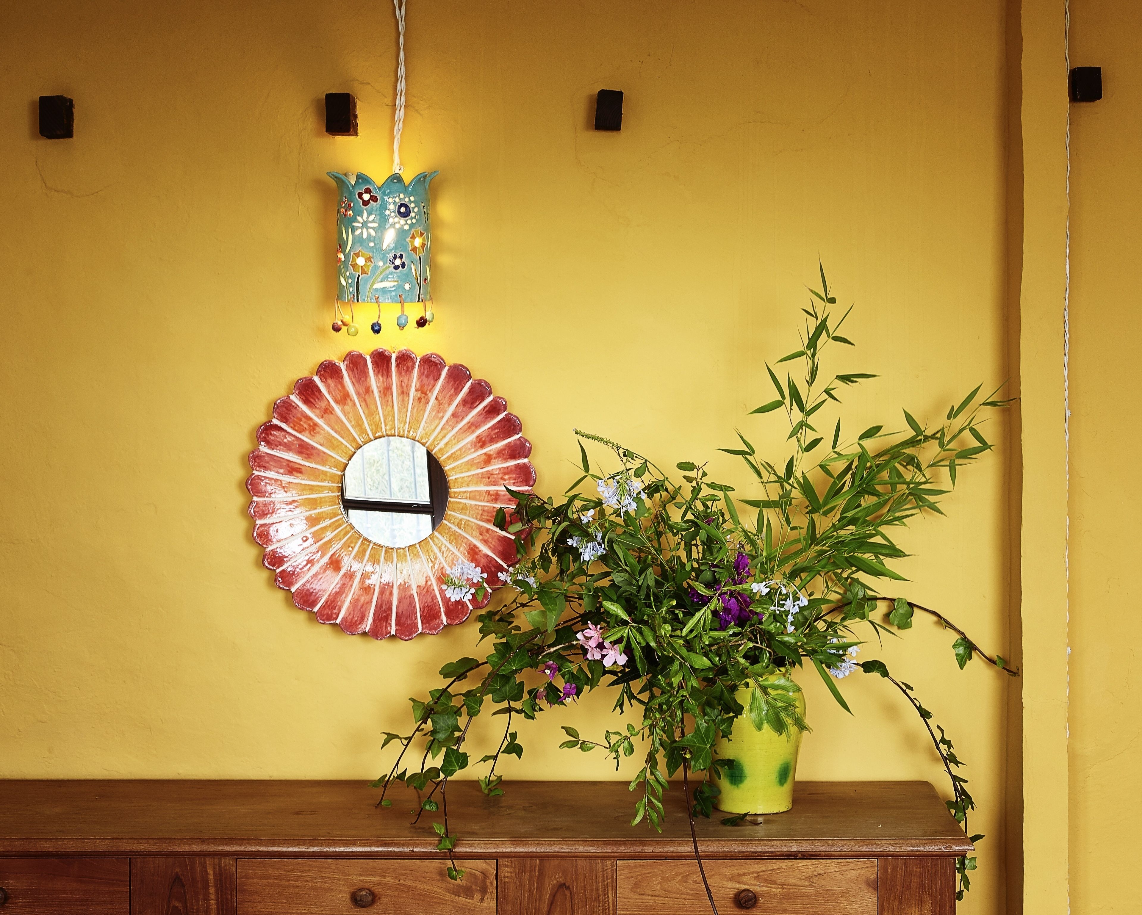 Een houten dressoir met een spiegel, een decoratief bord en een bloeiende plant voor een gele muur.
