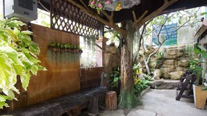 A covered outdoor seating area with a wooden bench, a stone wall, and various plants. - Kaya Place (Bangkok)