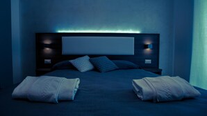 Uma cama bem arrumada com travesseiros e um cobertor, uma mesa de cabeceira com luminária, e uma luz na parede.
