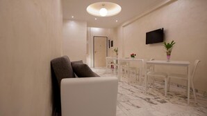 Quarto de hotel moderno com sofá branco, mesa de jantar com cadeiras, televisão de tela plana e uma planta em vaso.
