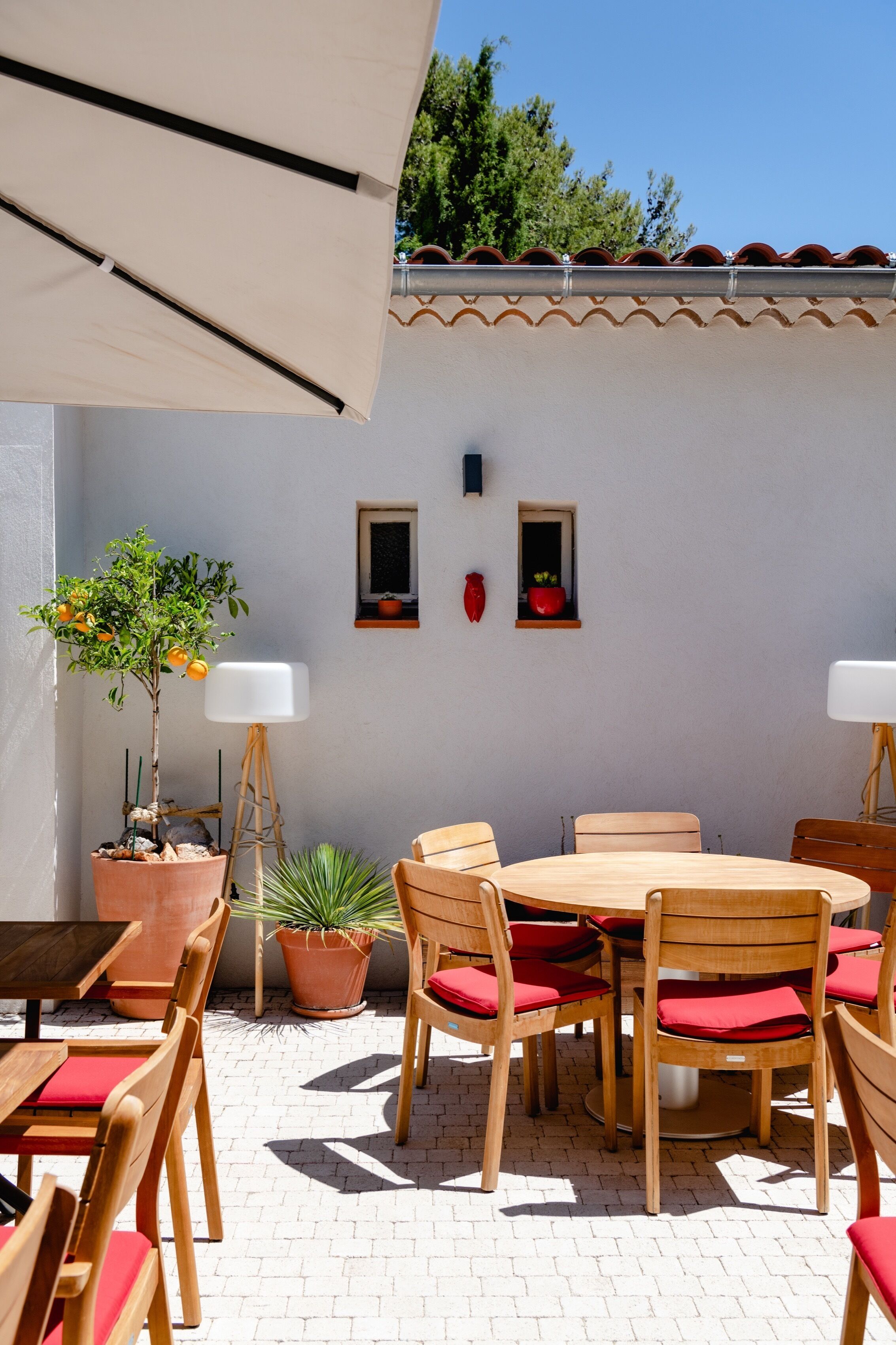 An outdoor patio with a round wooden table and chairs, surrounded by potted plants and a white wall with decorative elements.