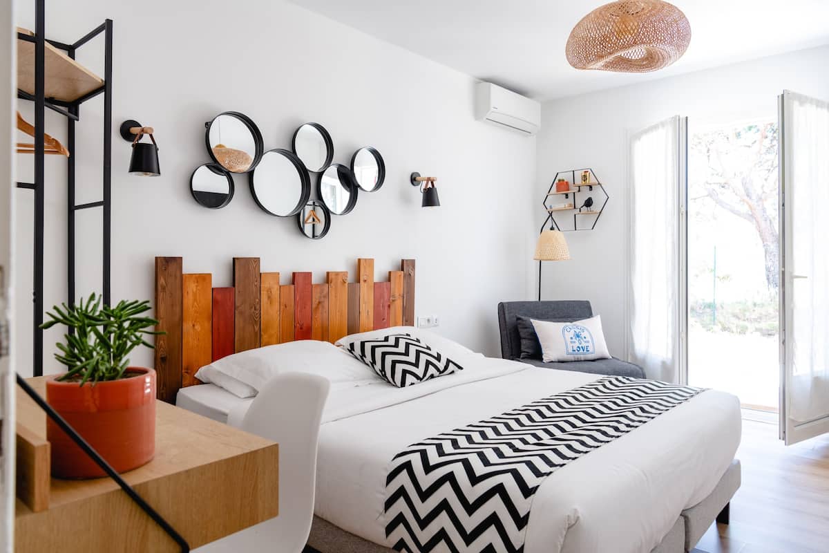 A modern bedroom with a bed, bedside table, potted plant, wall-mounted mirrors, and a view of the outdoors.