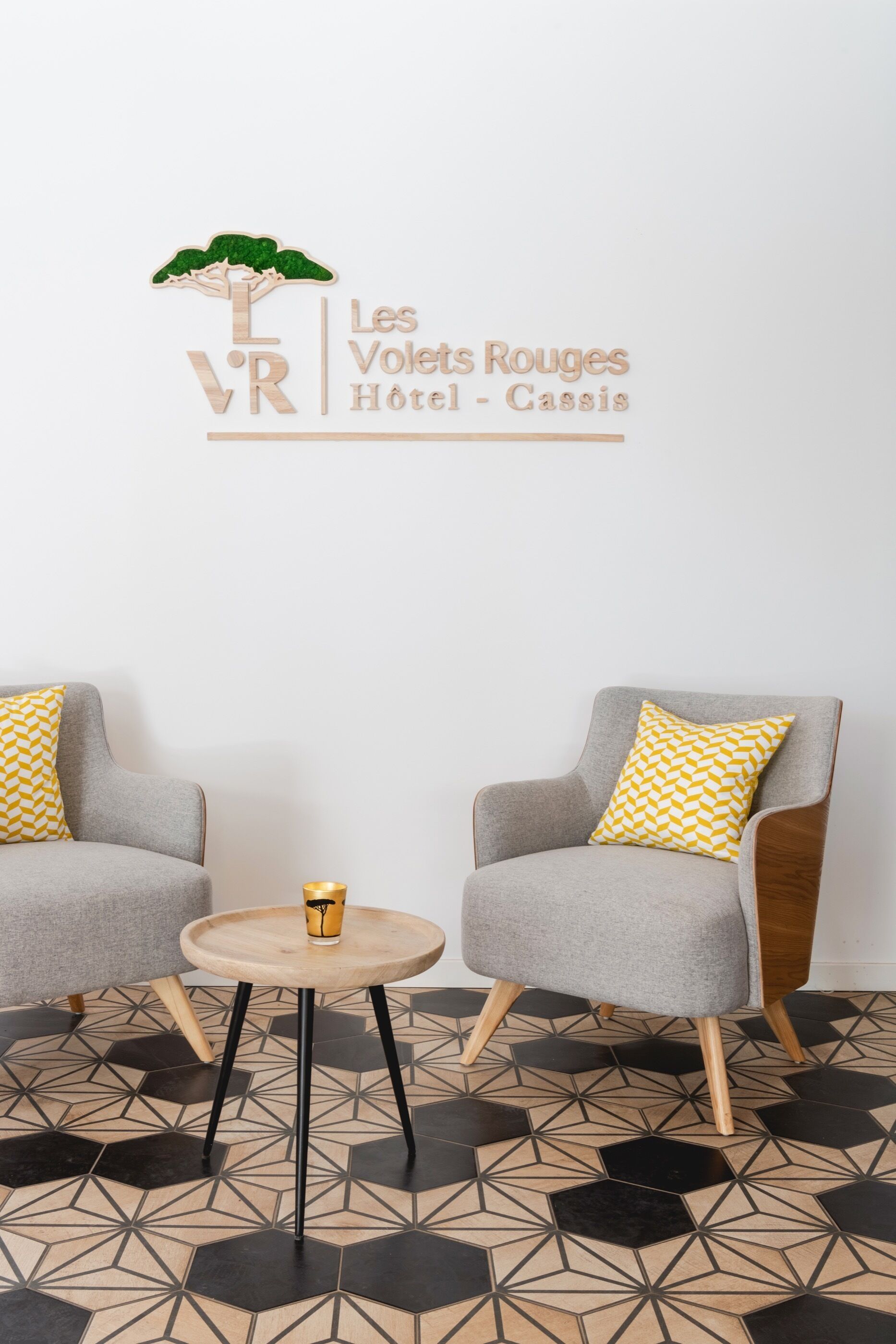 Une chambre d’hôtel moderne avec deux fauteuils gris, une petite table ronde et un sol orné d’un motif géométrique.