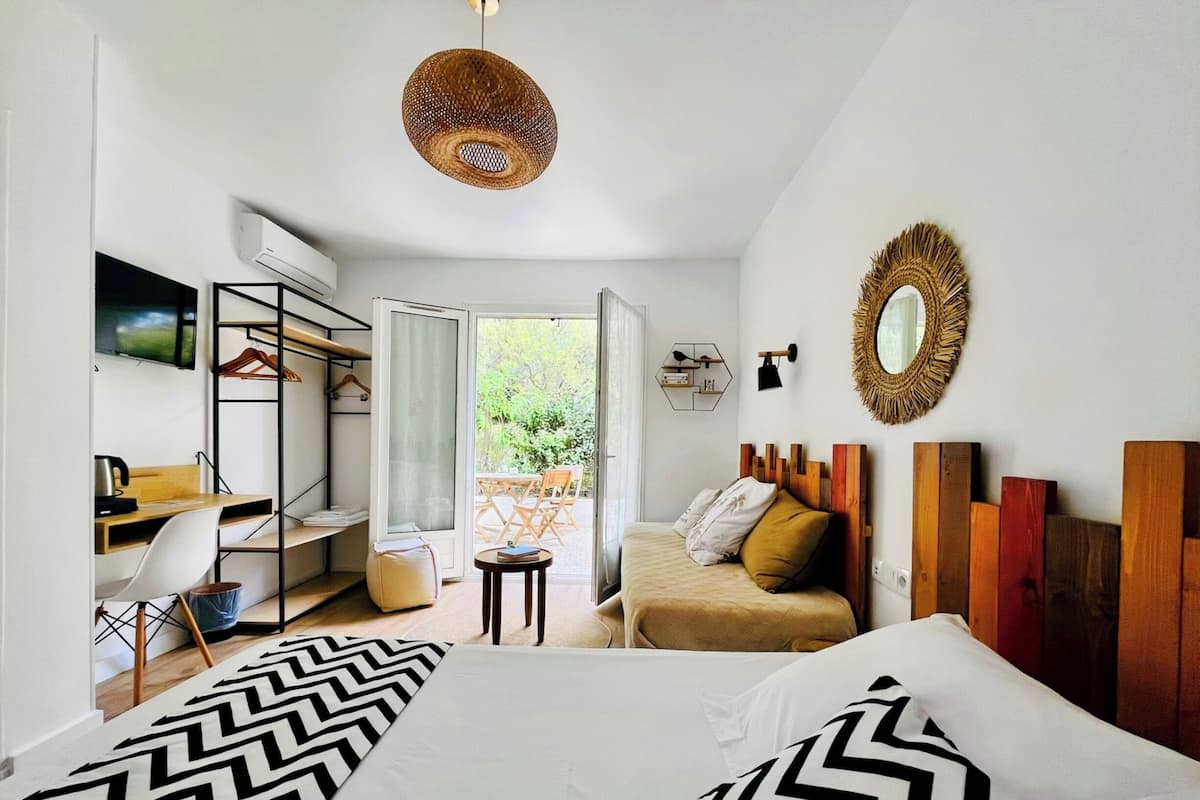 A modern bedroom with a bed, a desk, a chair, and a small table. There is a window with a view of greenery outside.