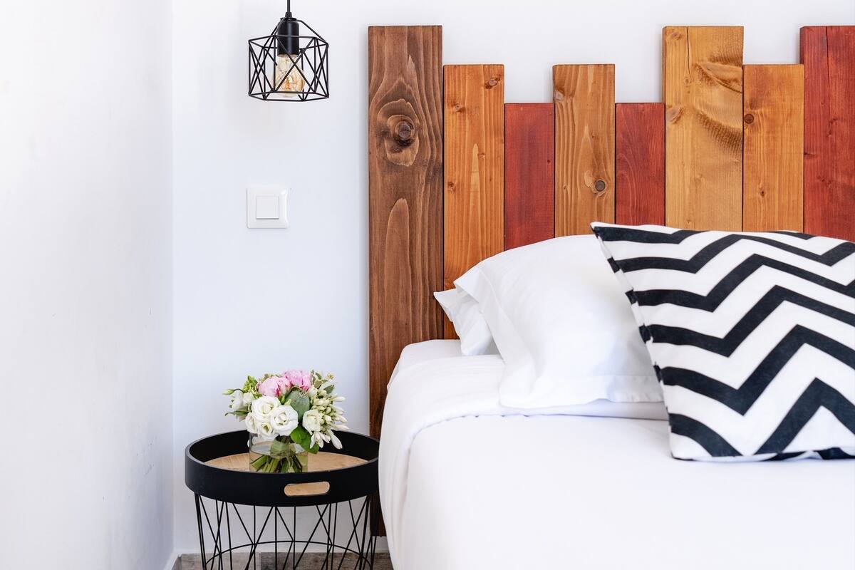 A neatly arranged bedroom with a wooden headboard, a bedside table with a geometric lamp, and a bed with a black and white chevron-patterned bedspread.