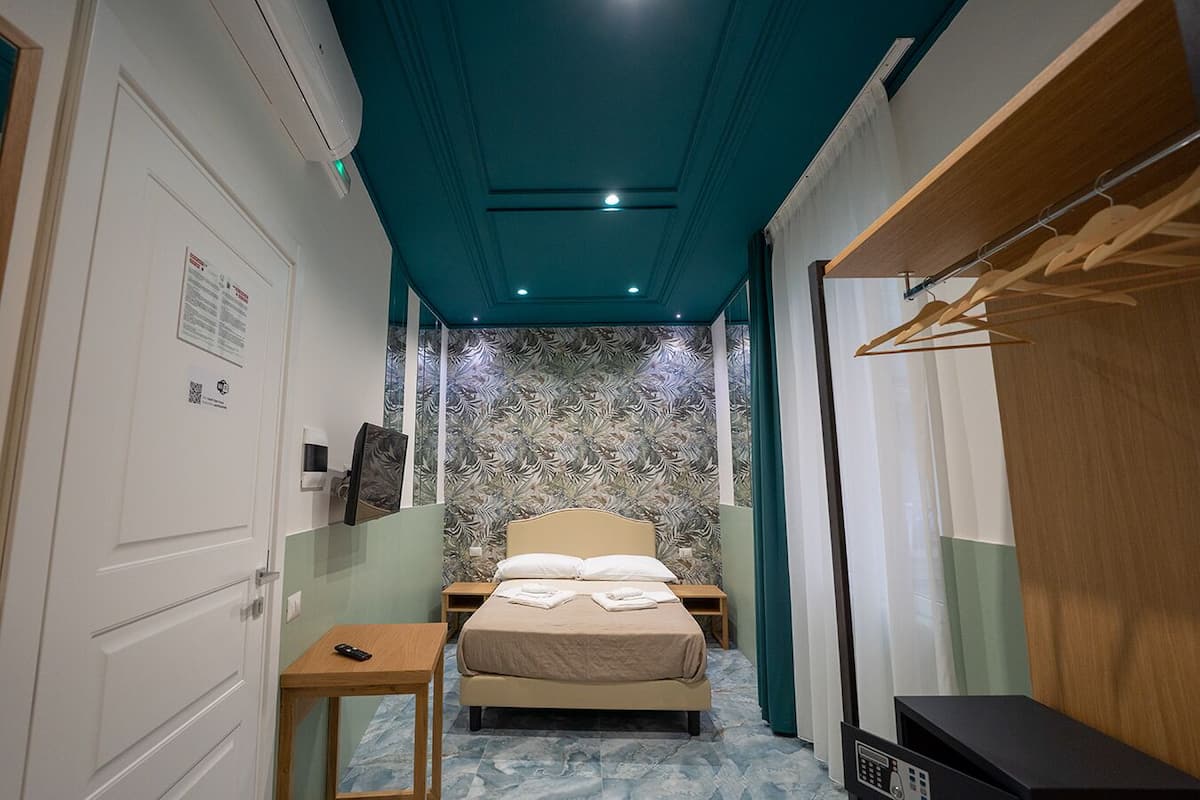 A narrow hotel room with a bed, a bedside table, a small table, a wardrobe, and a mirror.
