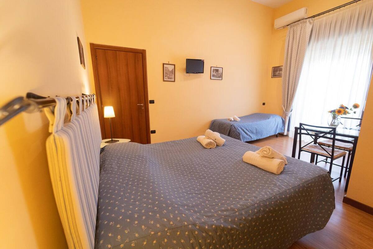 A hotel room with two beds, a desk, and a chair. There is a door, a wall-mounted TV, and a window with curtains.