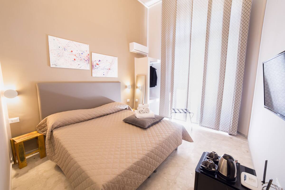 A bedroom with a large bed, a bedside table, a small wooden stool, a mirror, and a window with curtains.