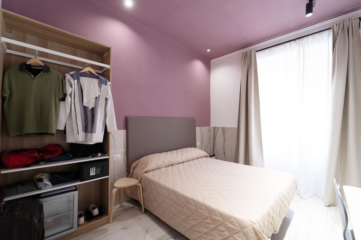 A bedroom with a bed, a wardrobe, a shelf with clothes, and a small table.