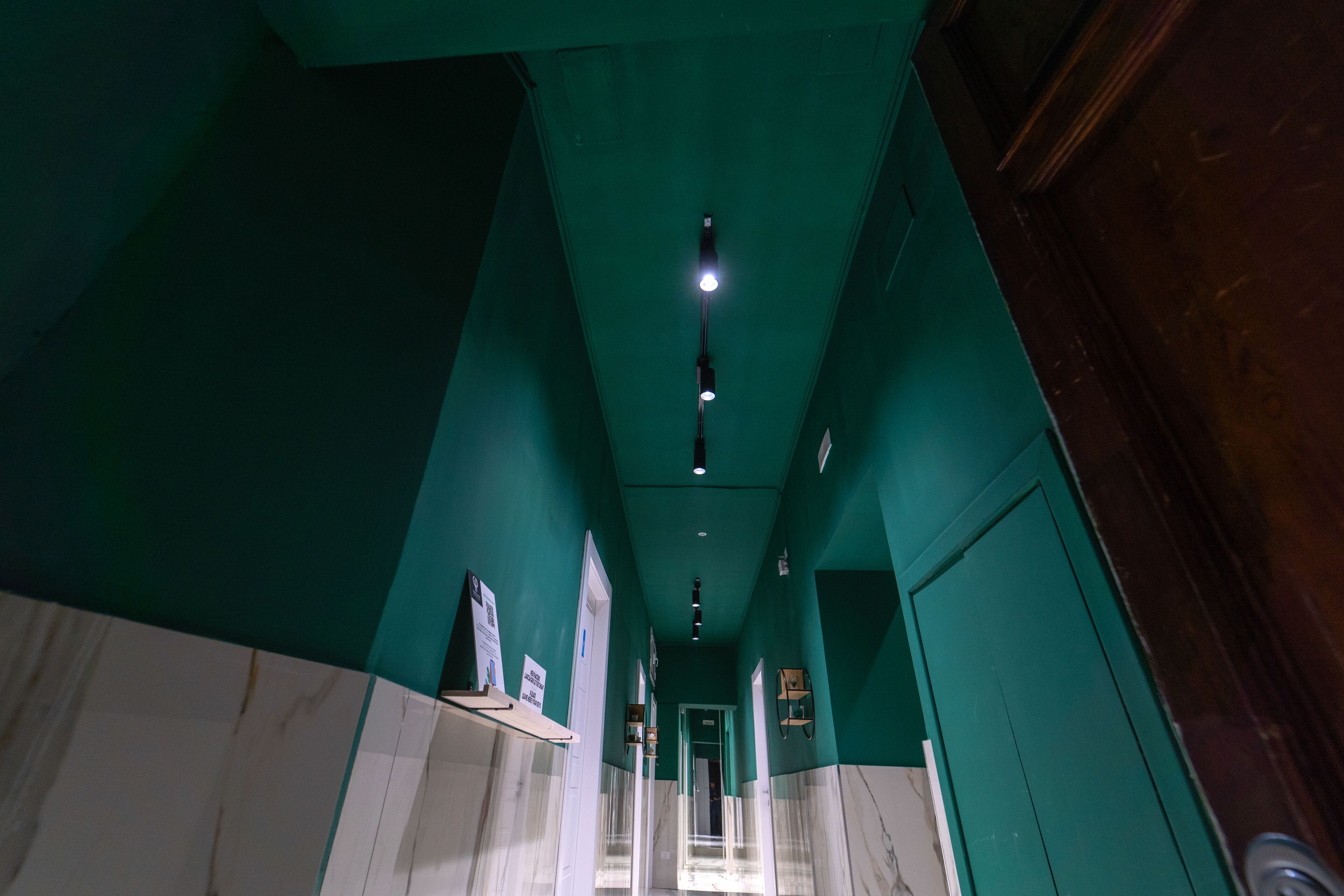 Un pasillo con paredes verdes, suelo de mármol blanco e iluminación empotrada en el techo.