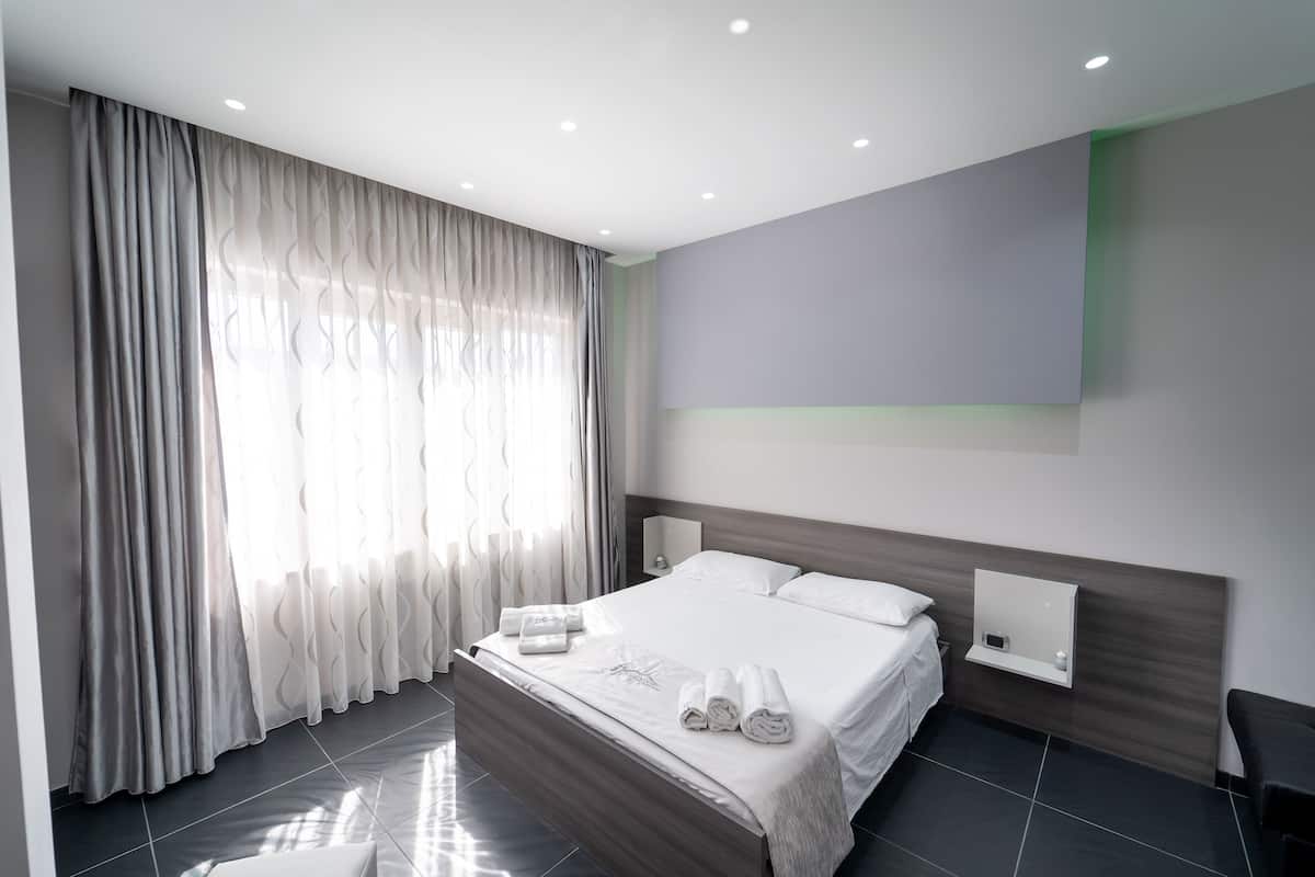 A modern hotel room with a large bed, a bedside table, a wall-mounted TV, and a window with sheer curtains.