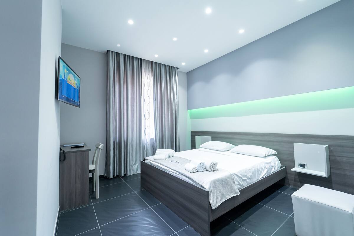 A modern hotel room with a neatly made bed, a desk with a chair, and a bedside table.