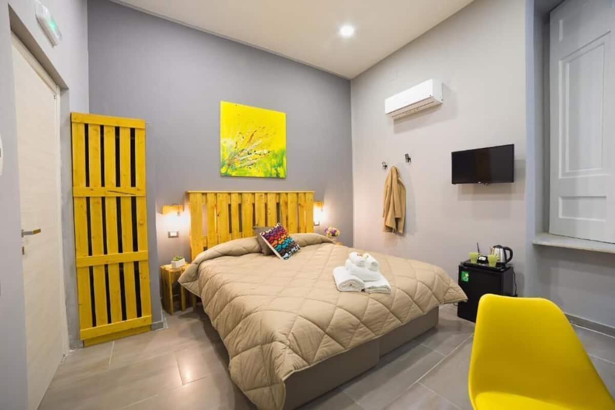 A bedroom with a bed, a yellow chair, a TV, and a painting on the wall.