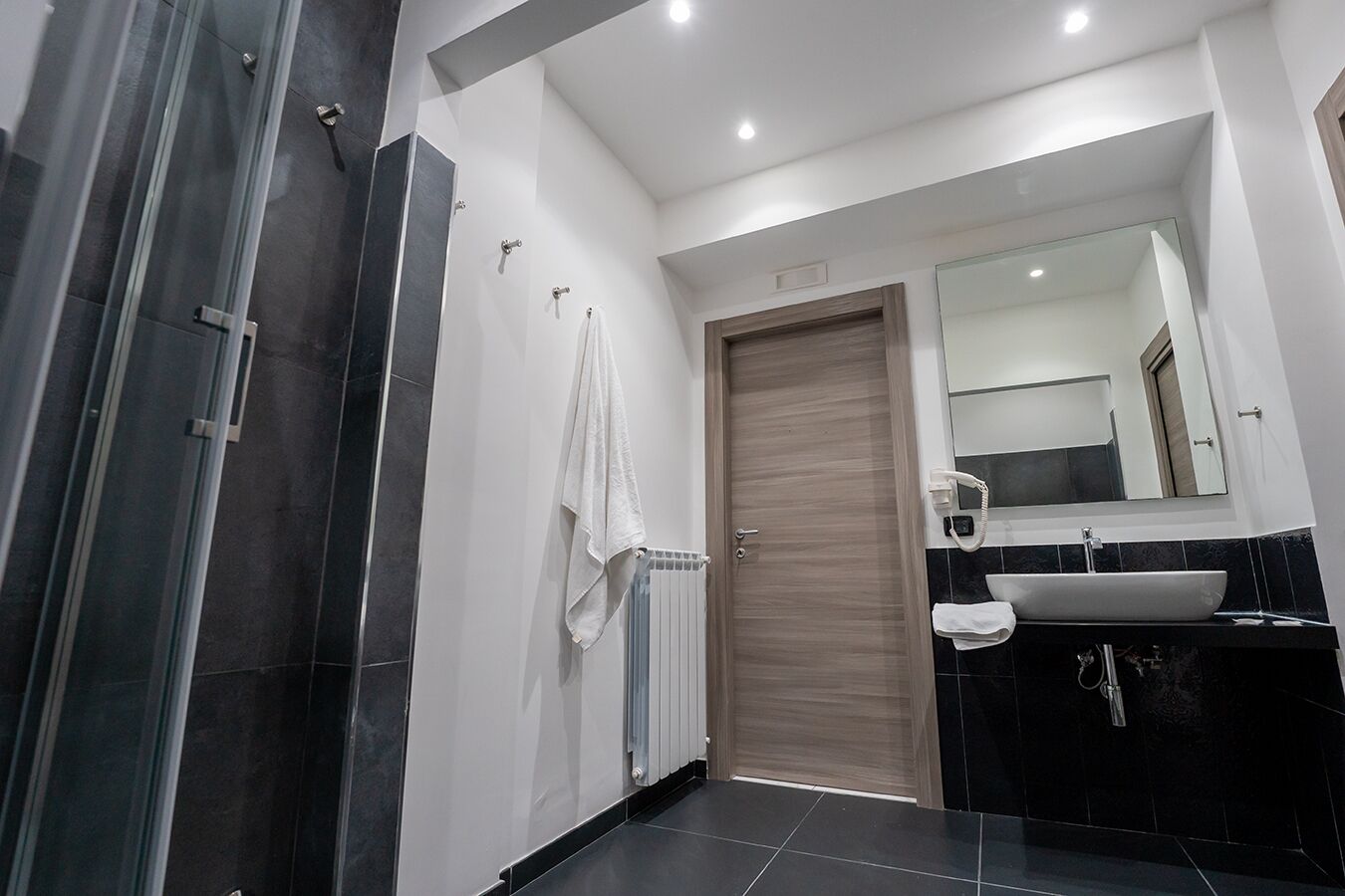 A modern bathroom with a glass shower enclosure, a wooden door, a white towel, a sink with a mirror, and black tiled flooring.