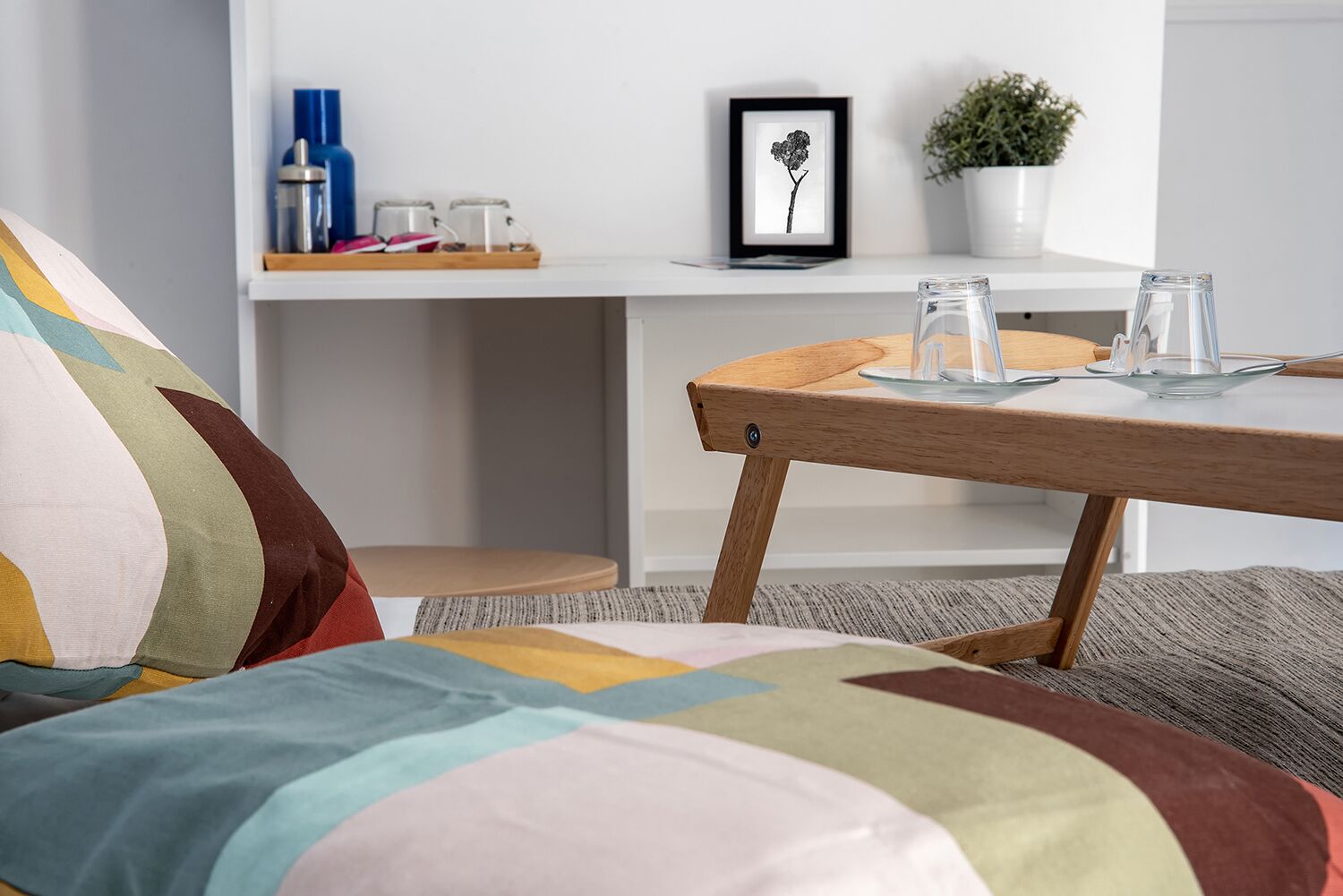 Une pièce chaleureuse avec une couverture de lit colorée et matelassée, une petite table en bois sur laquelle sont posés un pichet en verre et des verres, une étagère blanche ornée d’objets décoratifs et un tableau représentant une fleur.