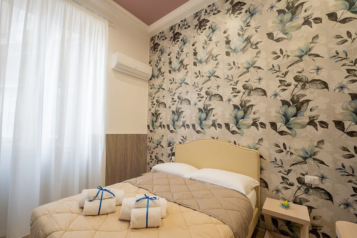 A hotel room with a bed, floral wallpaper, an air conditioning unit, and a bedside table with a glass.