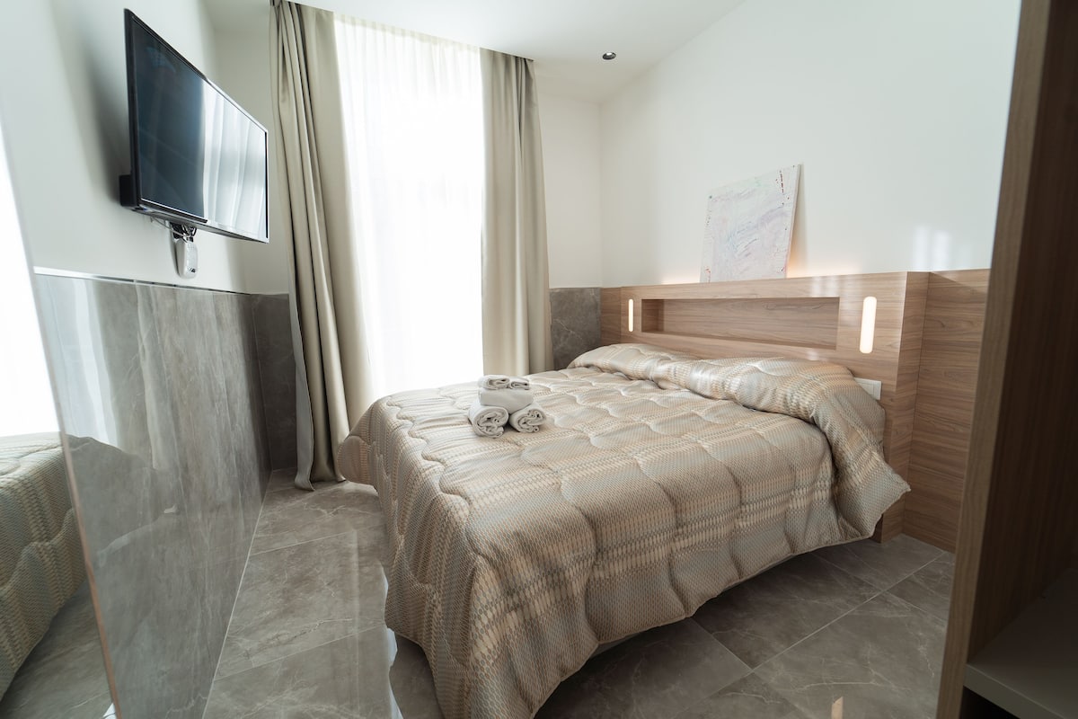 A modern hotel room with a large bed, a flat-screen TV mounted on the wall, and a large window with curtains.