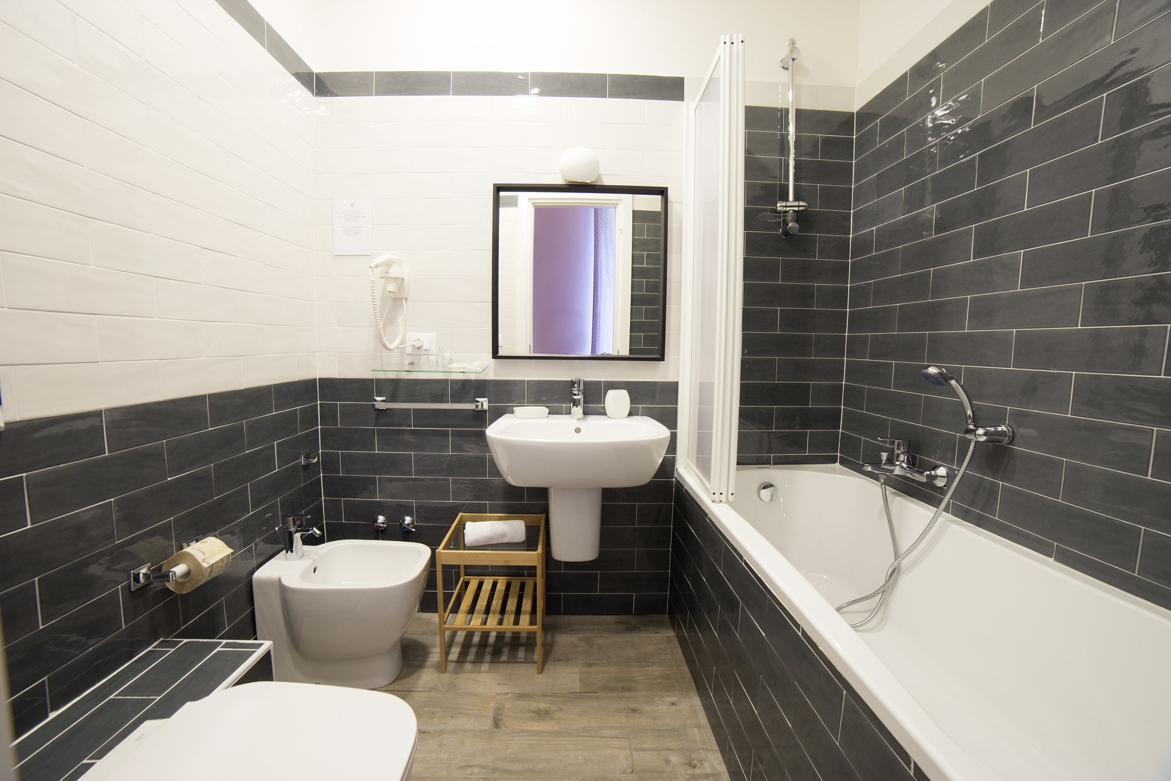 Ein modernes Badezimmer mit einem weißen Waschbecken, einer Toilette, einem Bidet, einem hölzernen Hocker und einer Badewanne mit Duschkopf.