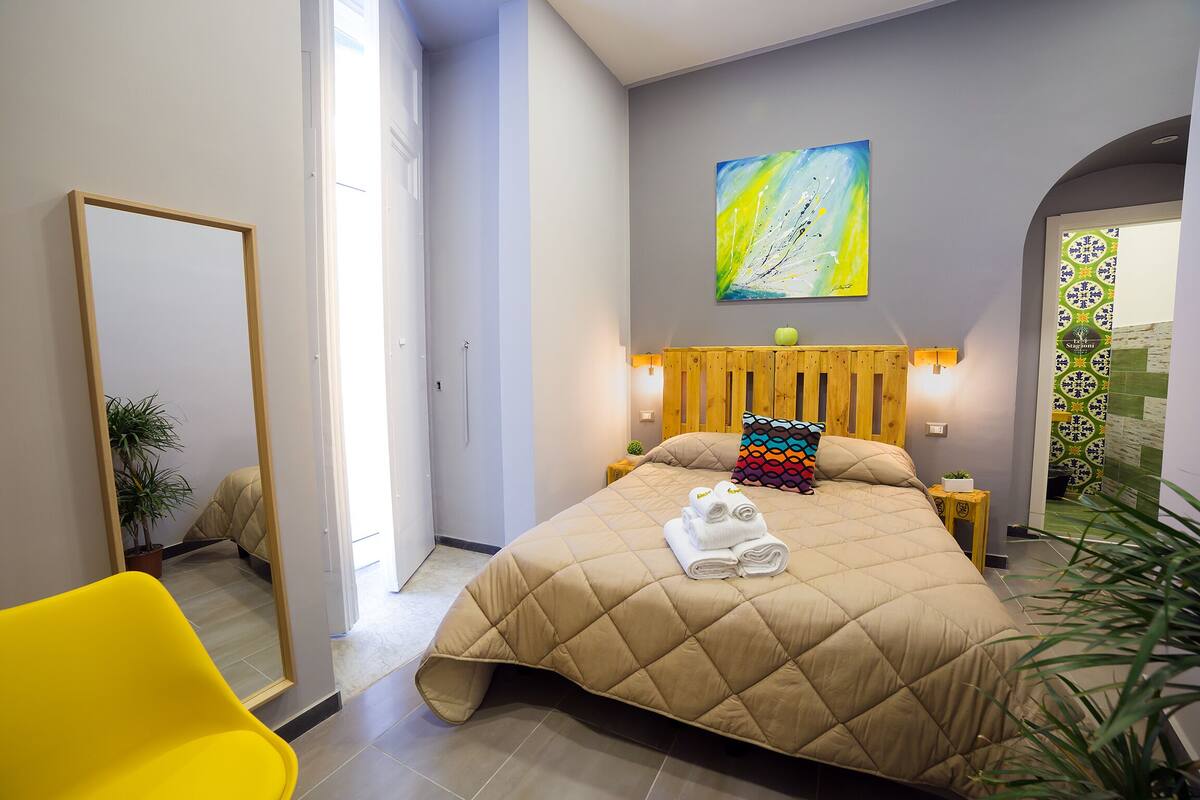 A bedroom with a large bed, a mirror, a yellow chair, and a potted plant.