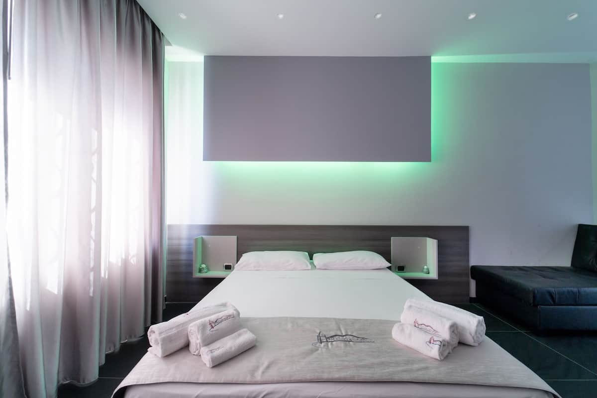 A modern hotel room with a large bed, a flat-screen TV mounted above it, and a bedside table with a lamp.