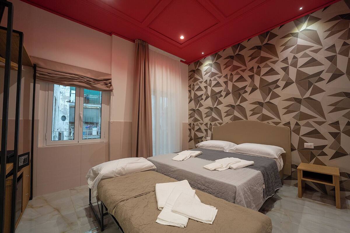Ein Hotelzimmer mit zwei Betten, einem Fenster mit Blick, einer Wand mit geometrischem Muster und einer roten Decke.