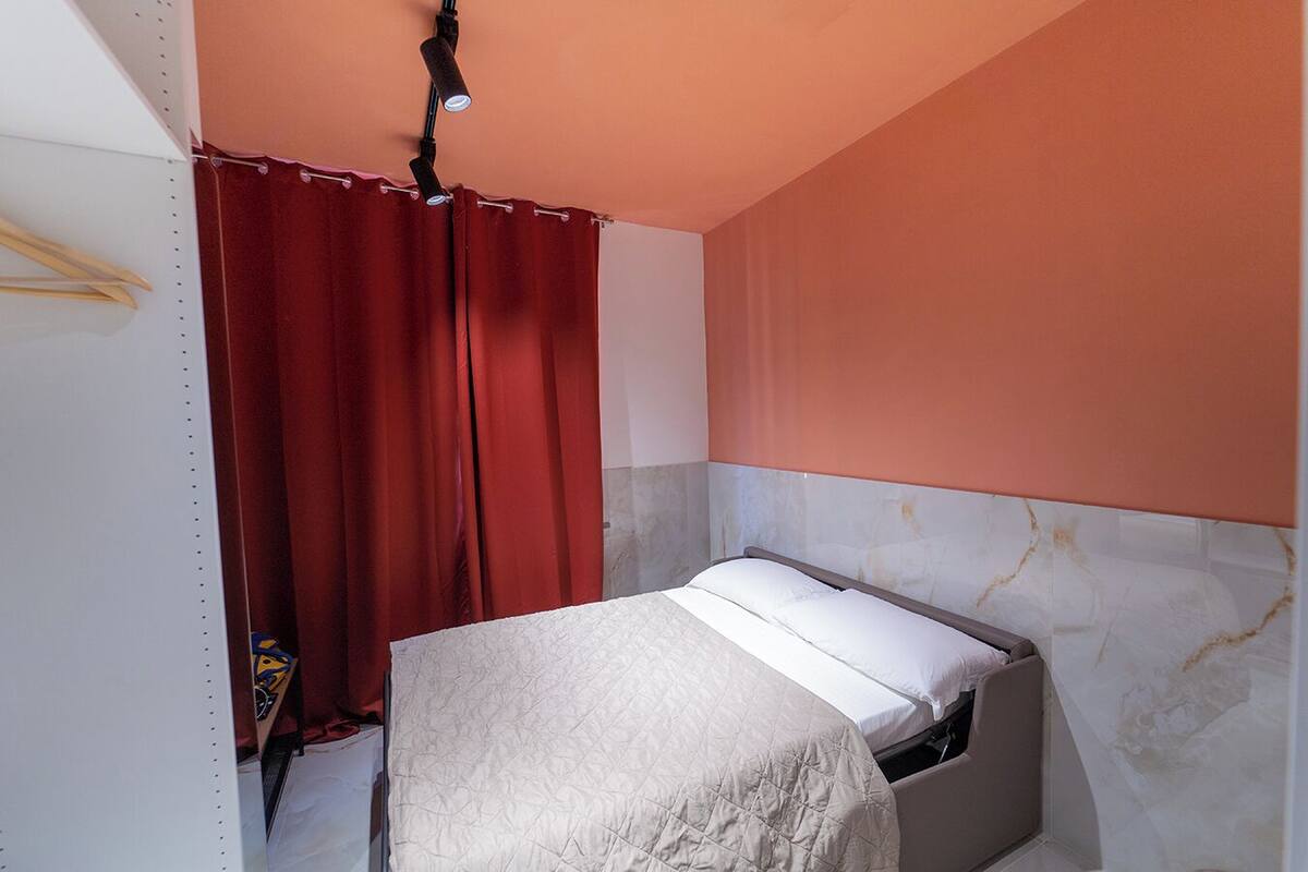 Ein kleines Zimmer mit einem Einzelbett, einem schlichten Bett, einem roten Vorhang und einer weißen Wand mit Marmorstruktur.