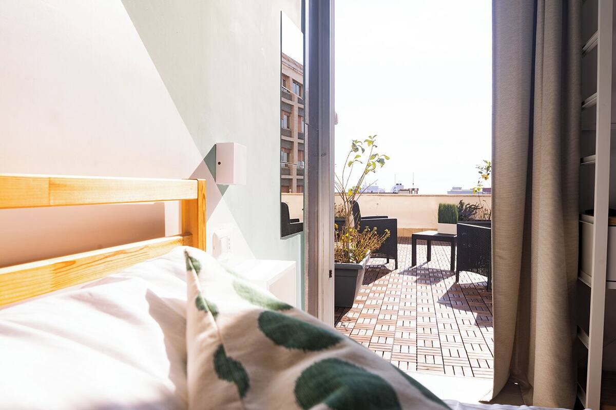 Ein Schlafzimmer mit Bett, Fenster und Blick auf eine Terrasse mit Tisch und Stühlen.