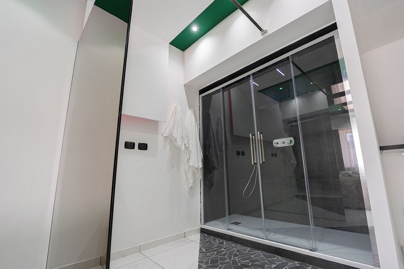 A modern bathroom with a glass shower enclosure, a white tiled floor, and a green accent on the ceiling.