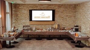 A breakfast buffet setup in a hotel room with a stone wall backdrop, a television screen displaying the hotel's name, and a variety of breakfast items on a long table. - Hotel Briliant (Cluj-Napoca)