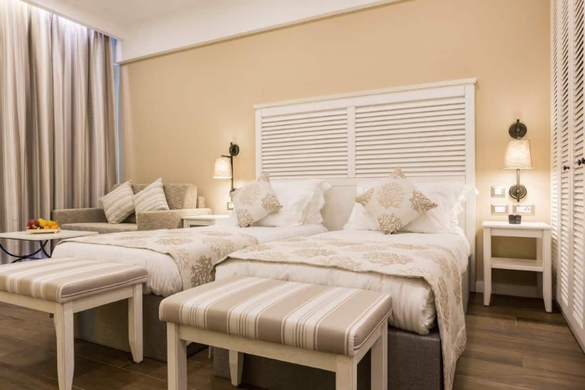 Ein Hotelzimmer mit einem großen Bett, zwei Nachttischen, einer Bank, einem kleinen Tisch mit Obst und einem Fenster mit Jalousien.