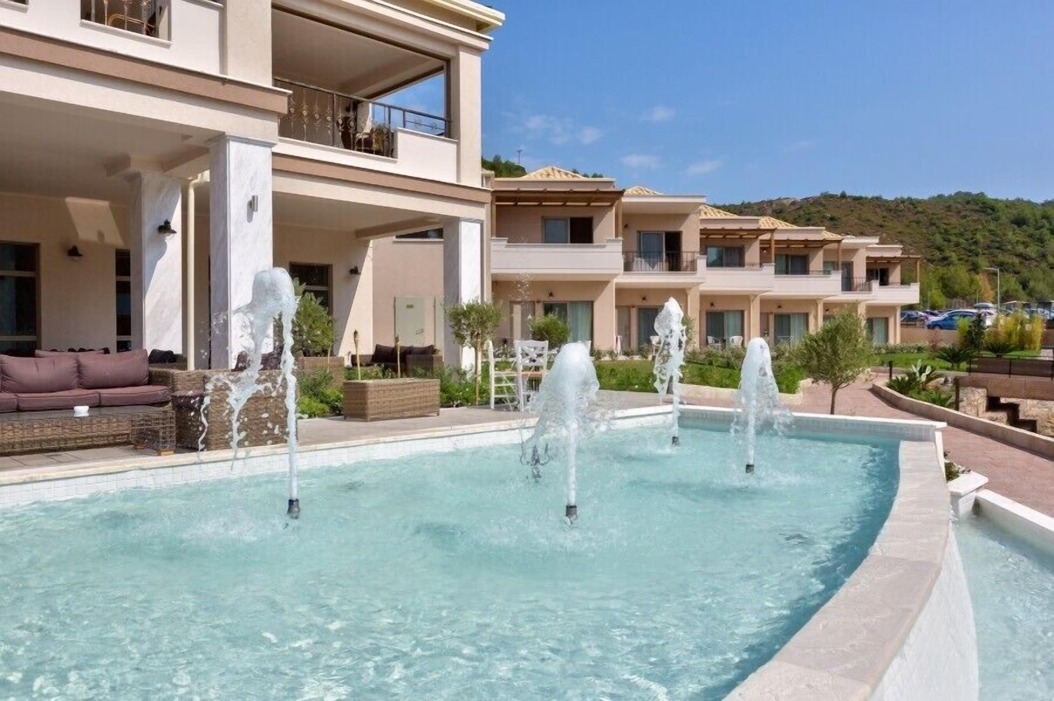 Una piscina con múltiples chorros de agua, rodeada por un edificio con balcones y vistas a un paisaje montañoso.