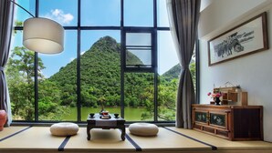 Una habitación con suelo de tatami, una mesa baja con un jarrón y una tetera, un ventanal con vistas a una montaña verde y un lago, y un armario de madera con un cuadro enmarcado.