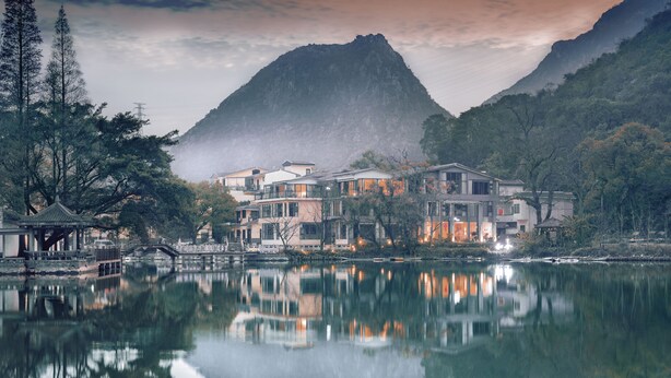 Un hotel frente al lago con arquitectura tradicional, su reflejo en el agua y montañas al fondo.