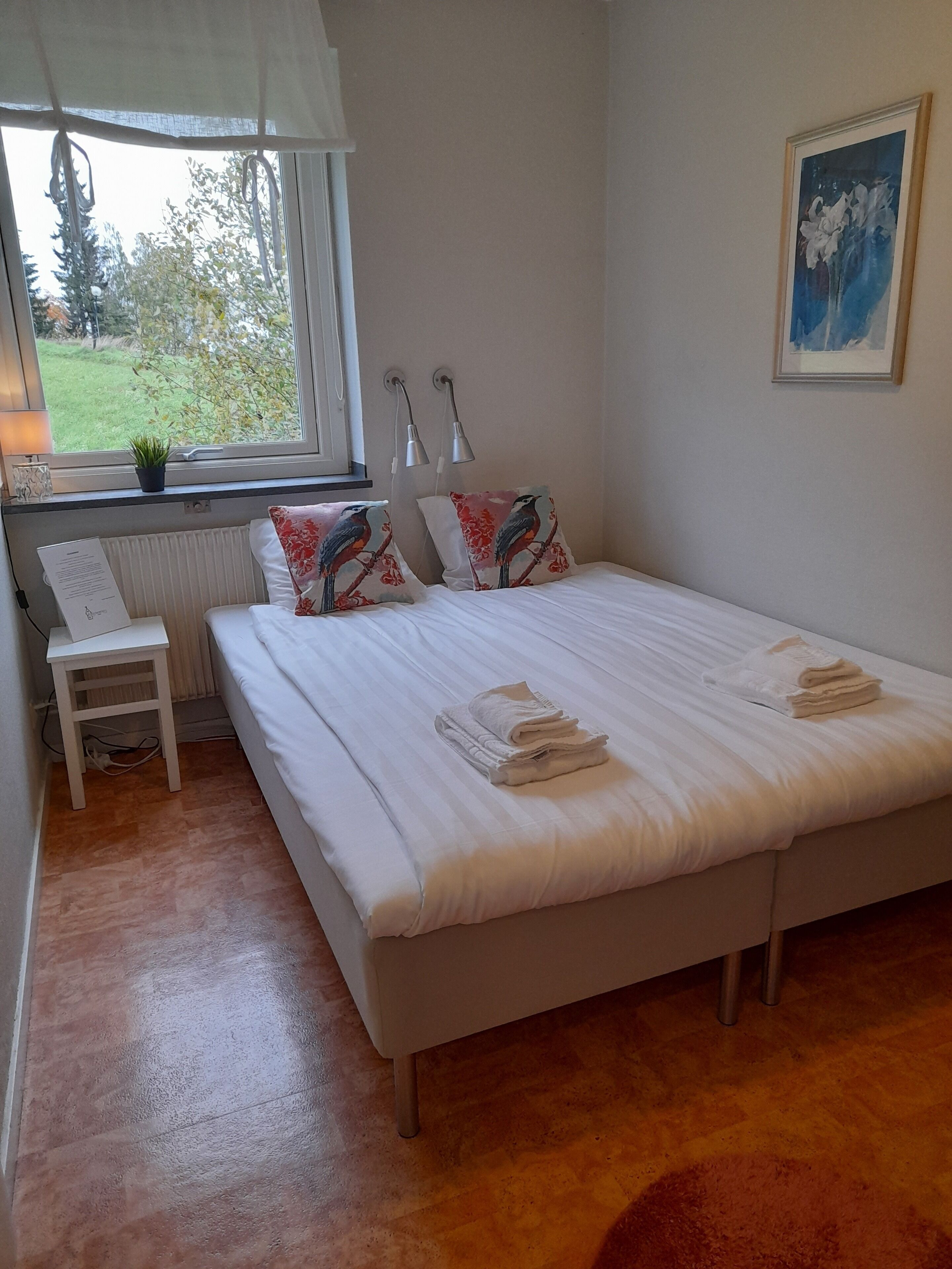 Un dormitorio con cama, dos almohadas con diseños de pájaros, una mesita de noche, una lámpara, una ventana con vistas a zonas verdes y un cuadro en la pared.