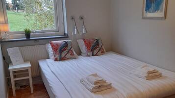Un dormitorio con cama, dos almohadas con diseños de pájaros, una mesita de noche, una lámpara, una ventana con vistas a zonas verdes y un cuadro en la pared.