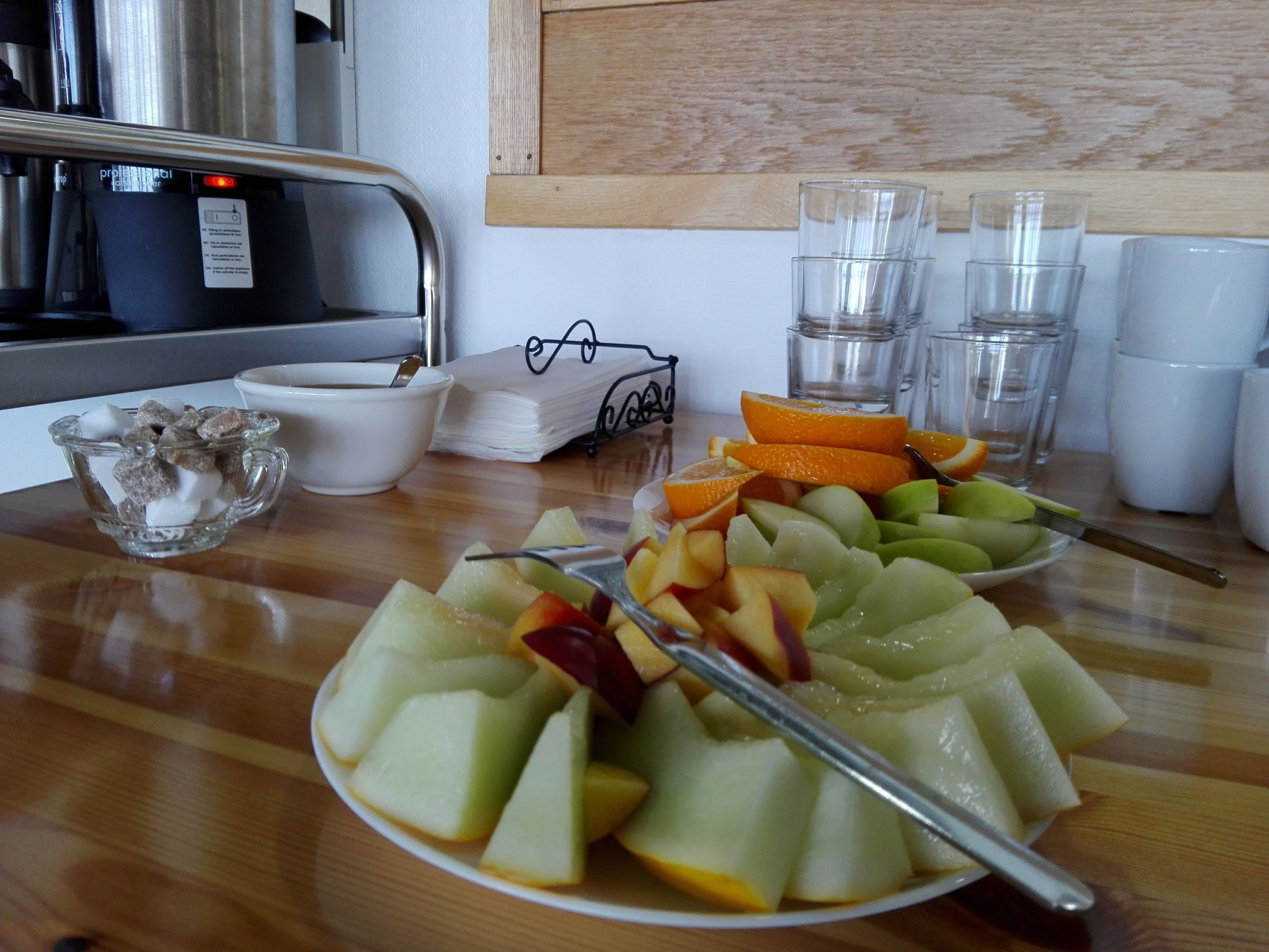 A breakfast table with a plate of fruit, a cup of coffee, and a small bowl of sugar cubes.