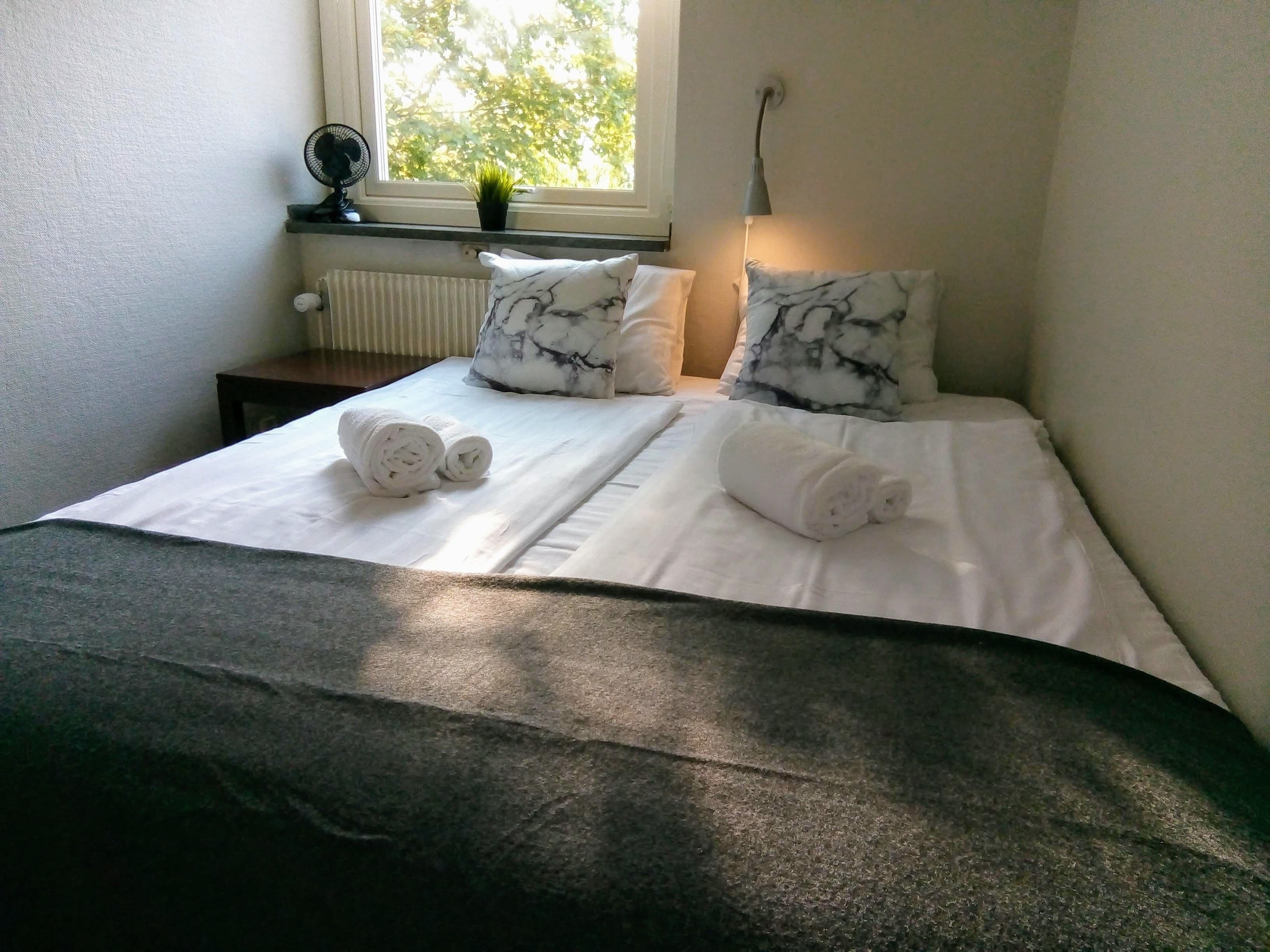A neatly made bed with white linens and marble-patterned pillows, a small table with a fan and a plant, and a window with a view of trees.