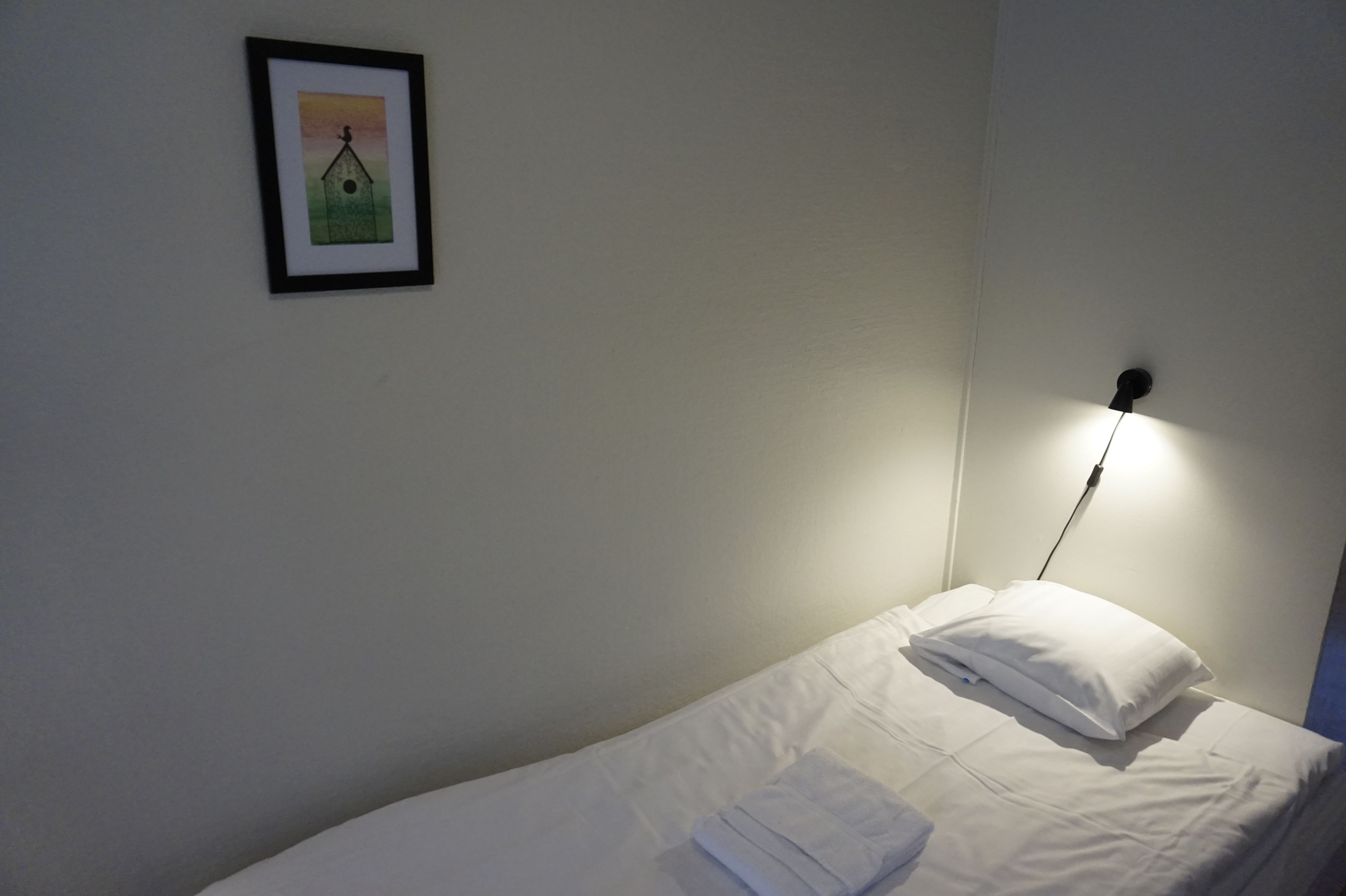 A single bed with white bedding and a folded towel, a framed picture on the wall, and a bedside lamp.