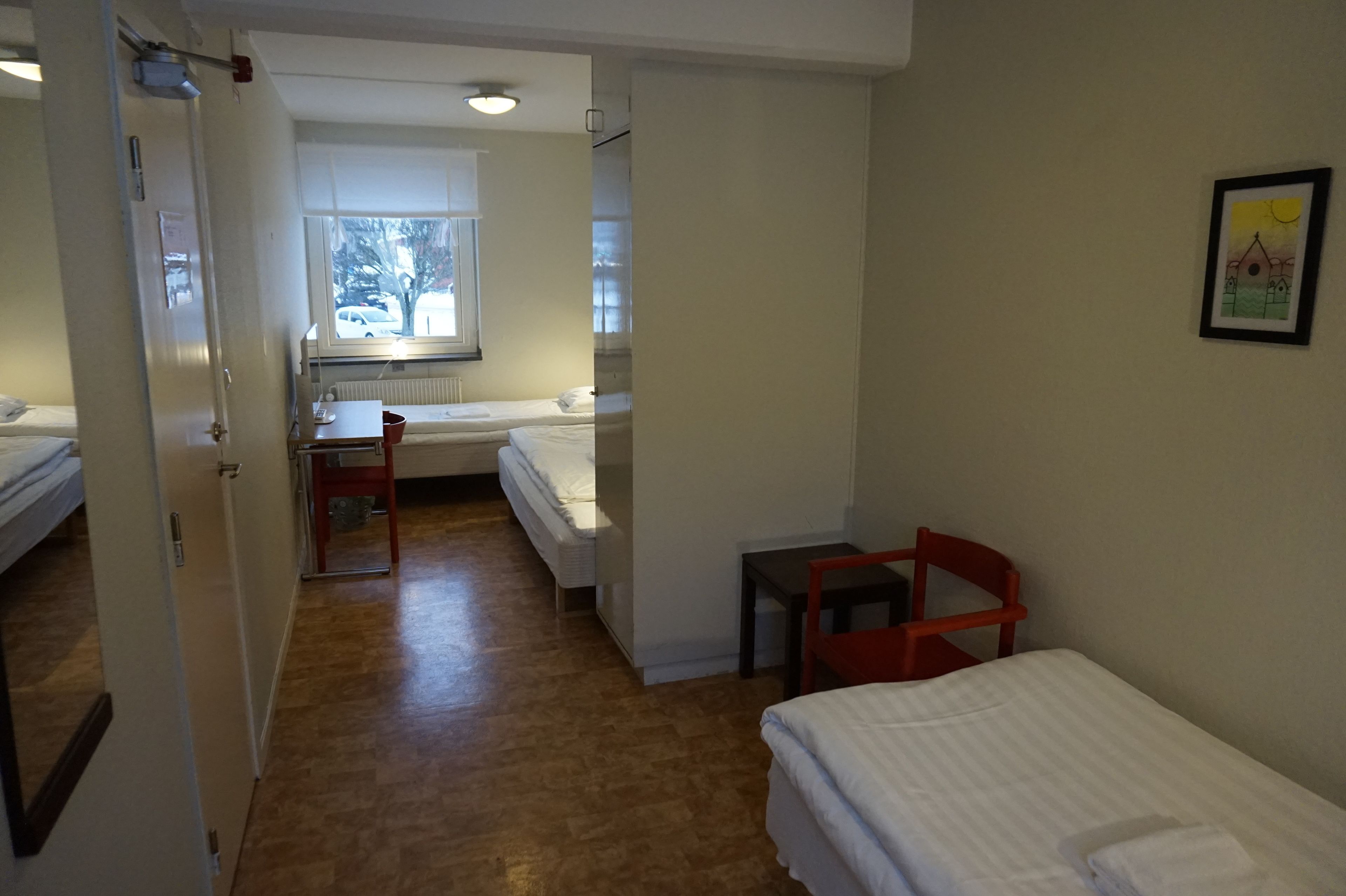 A room with two beds, a small table, and a chair. There is a window with a view of trees and a car outside.
