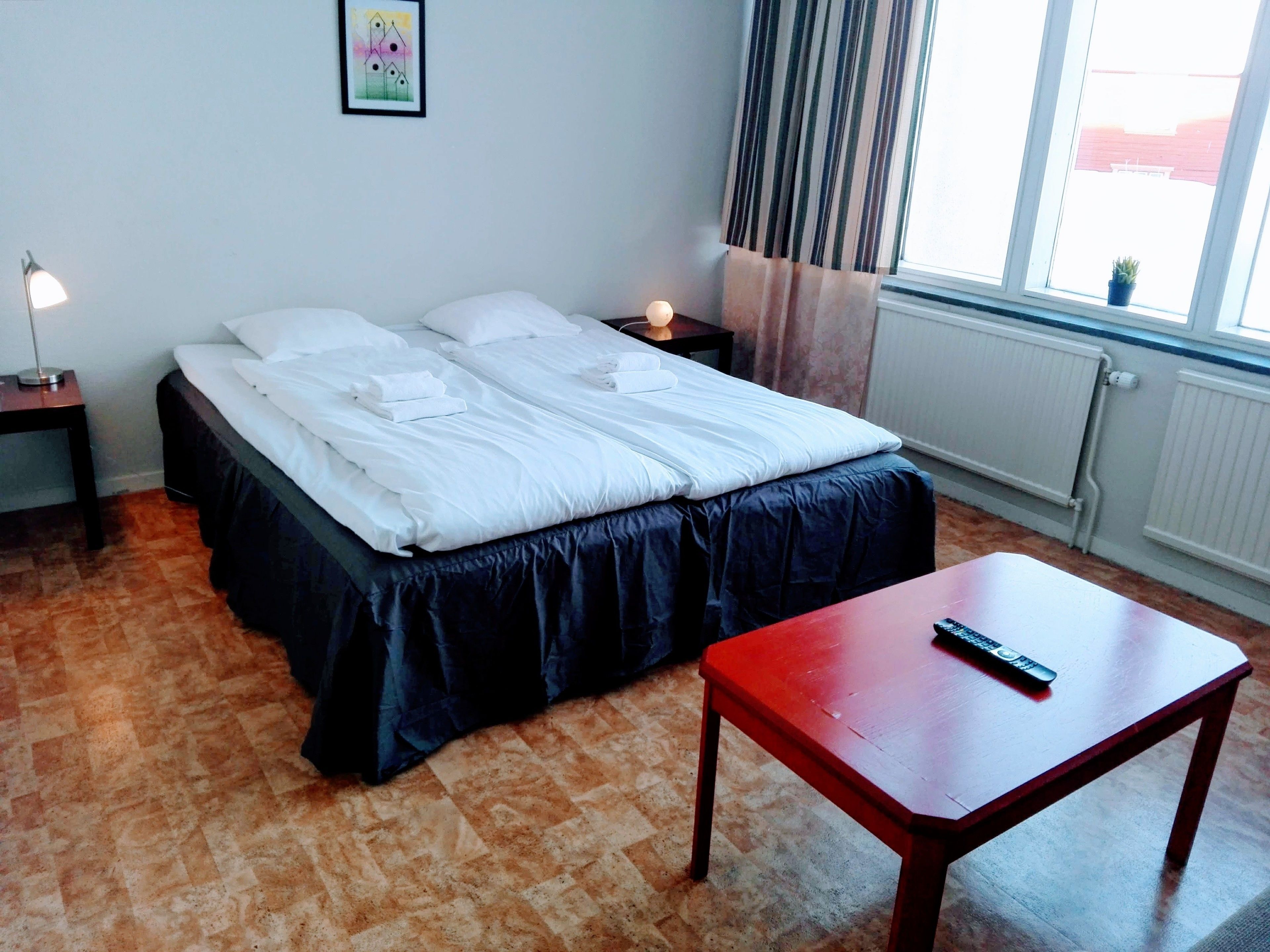 A neatly made bed with white linens, a wooden nightstand with a lamp, a small table with a remote control, and a window with curtains.
