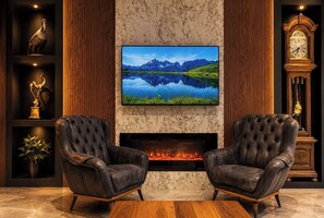 A room with a fireplace, two leather armchairs, a television mounted above it, and a wooden cabinet with decorative items. - Sezgin Hotel (Çaykara)