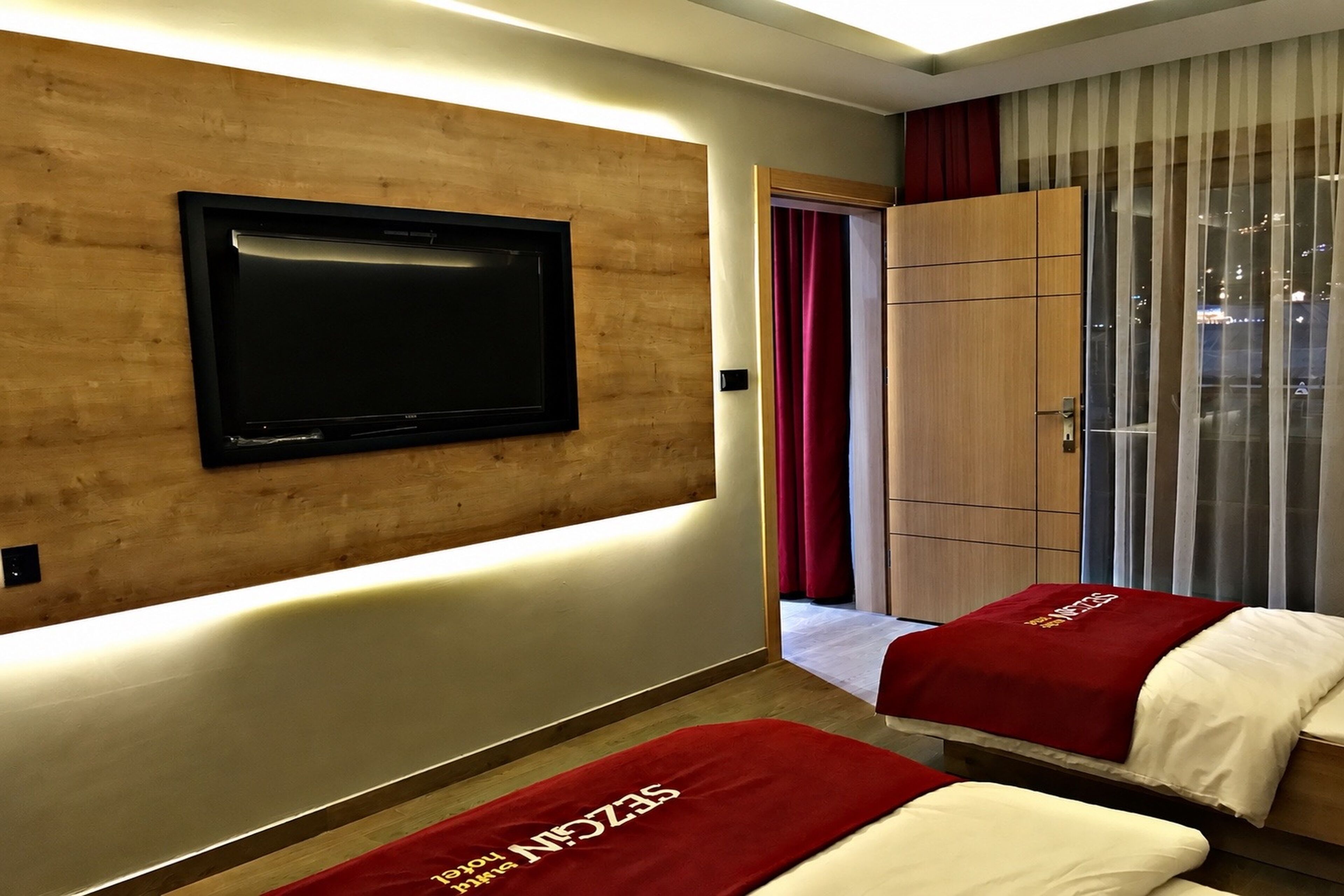 A hotel room with a flat-screen TV mounted on a wooden wall, a large window with curtains, and two beds with red bedspreads.