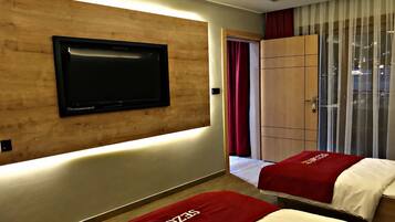 A hotel room with a flat-screen TV mounted on a wooden wall, a large window with curtains, and two beds with red bedspreads.