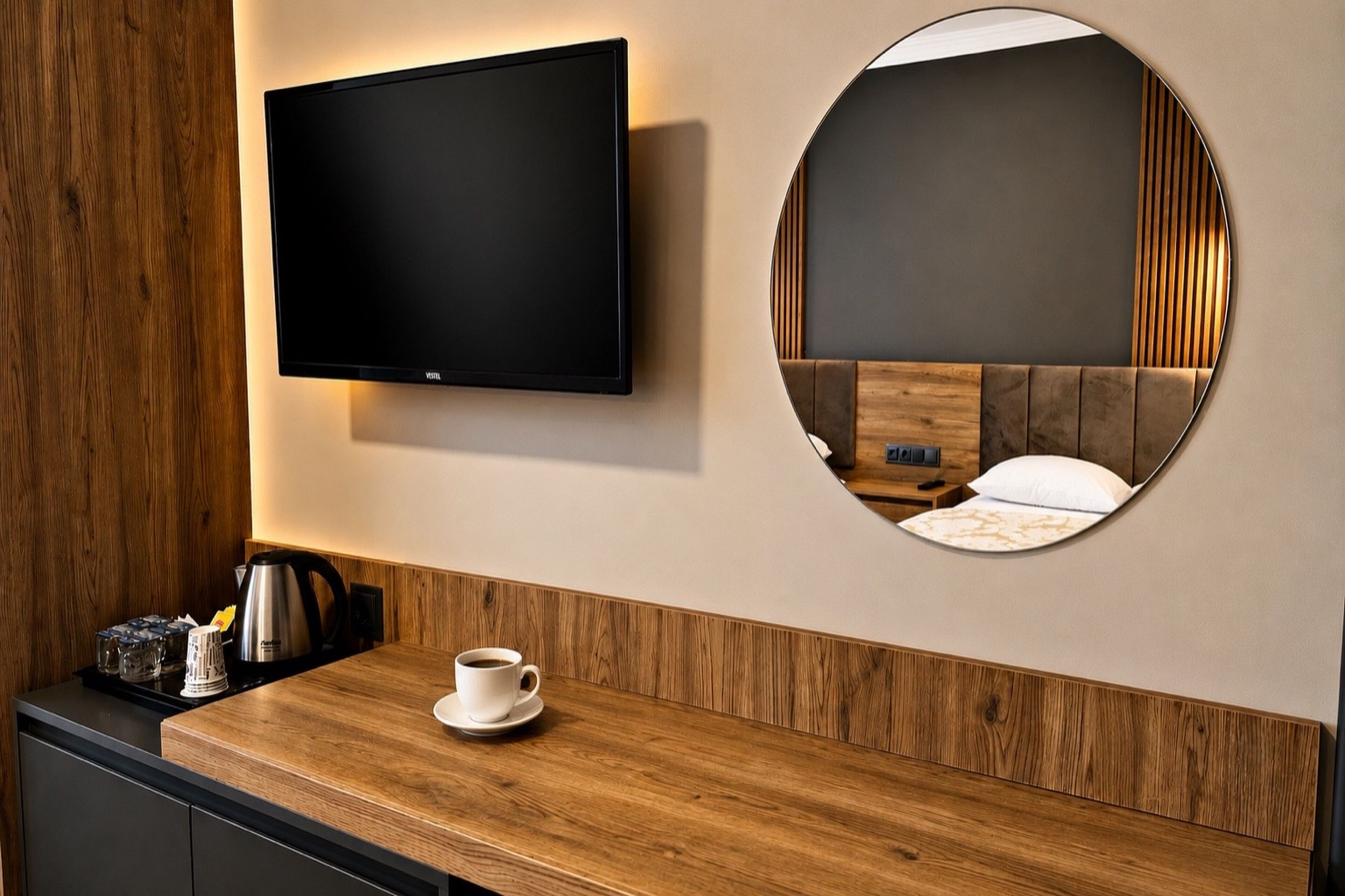 Quarto de hotel moderno com televisão de tela plana na parede, um espelho redondo refletindo a cama e um balcão de madeira com uma xícara e máquina de café.