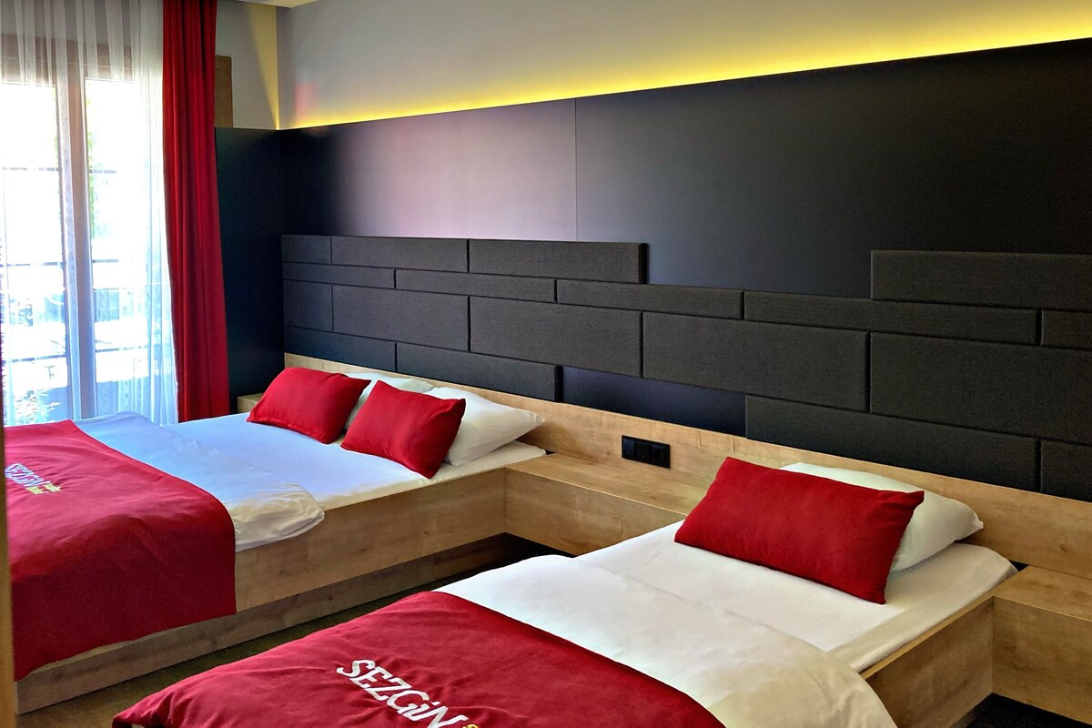 Two beds with red and white bedding, a dark headboard, and a window with red curtains.