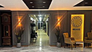 Un hall d’hôtel avec un sol en marbre, un comptoir d’accueil, deux plantes en pot, une horloge et un panneau indiquant « MEKKE ».