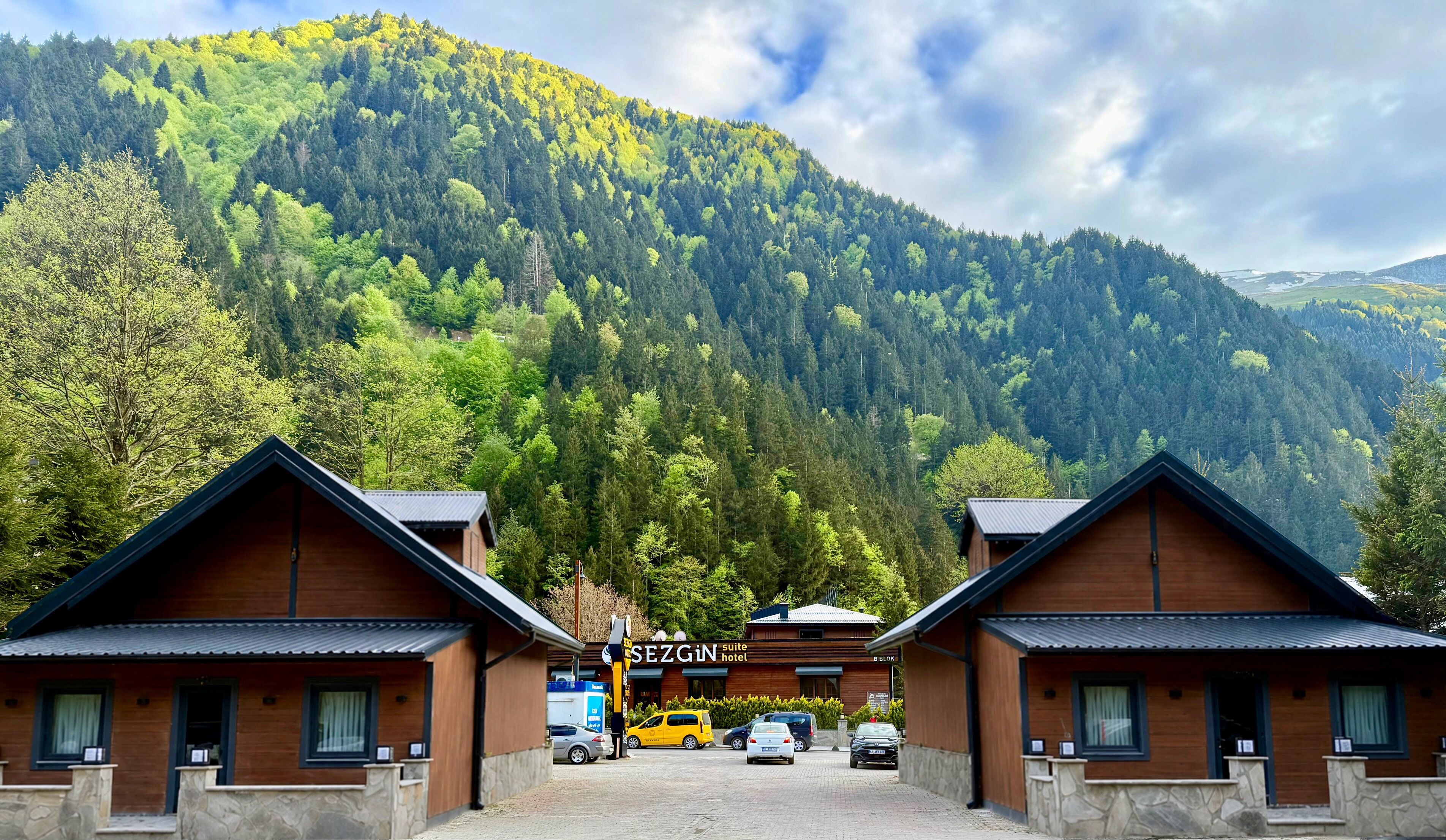 A hotel with a mountain backdrop, featuring a wooden facade, a signboard, and a parking area with cars.